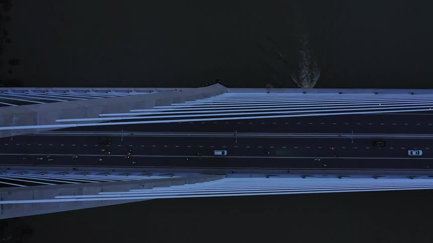 A topdown aerial view pans across a cablestayed bridge filled with evening traffic to reveal a boat passing through the dark river water underneath