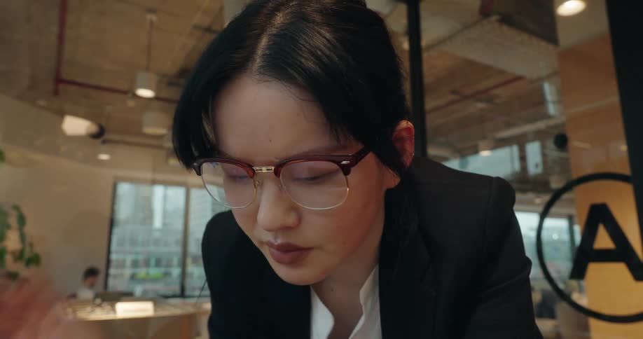 A woman in a black blazer and white shirt is seen working on a laptop in a modern office settingshe stops working for a moment to put on a pair of brown halfrimmed glasses before returning her attention to the screen