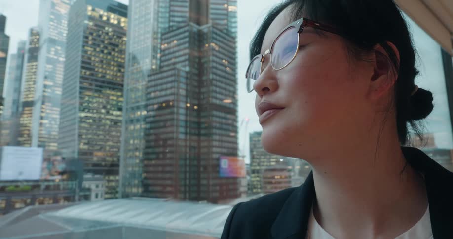 A closeup side view features a professional woman in glasses looking thoughtfully out of a large window at a dense cityscape dominated by tall modern skyscrapers