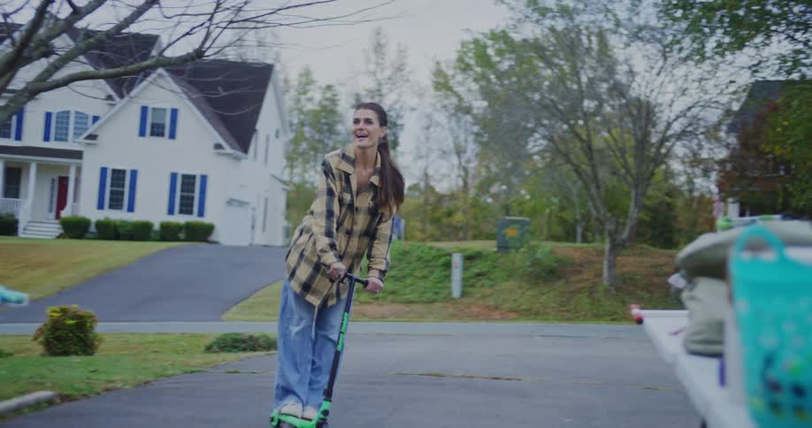 A woman rides a scooter down a suburban driveway as a yard sale table sits to the side
