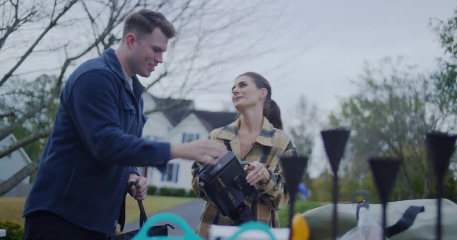 The video shows a man and a woman outdoors possibly setting up for a shoot or eventthe woman is holding a professional video camera and smiles as she hands it to the man who then lifts it to his shoulder ready to film