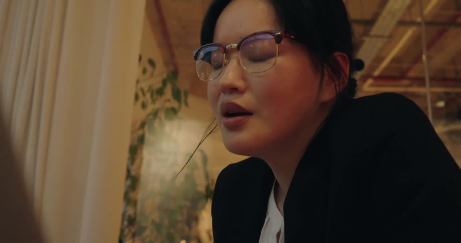 A woman wearing glasses talks while looking at a laptop screen in a warm office setting