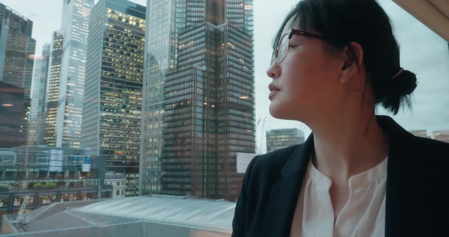 A businesswoman wearing glasses gazes out a window toward the city skyline