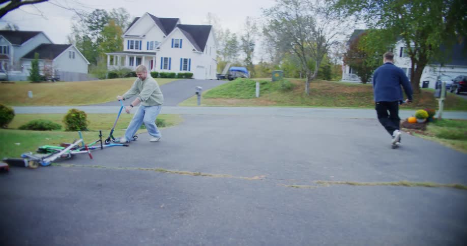 Two men in casual clothes ride scooters in a playful way down a suburban driveway passing fallen scooters and bikes near large white houses