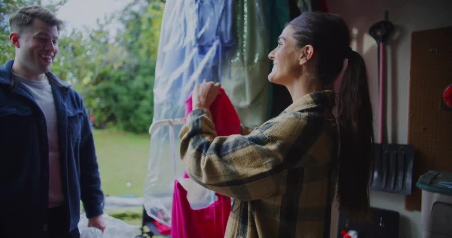 A smiling woman holds up a vibrant pink dress for a man to see while they sort through clothes in an open garage sharing a happy and nostalgic moment