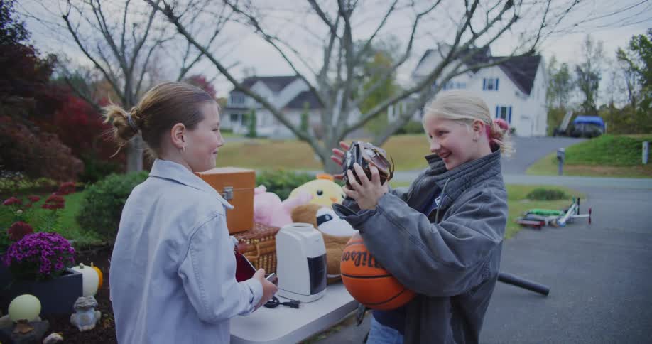 Two young girls are smiling and looking at a vintage camera over a table full of assorted items including a basketball and stuffed animals at an outdoor yard sale