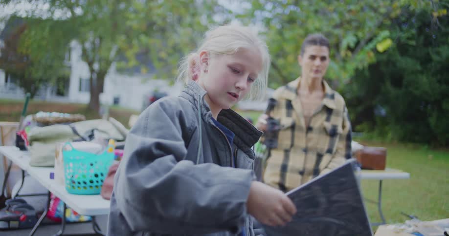 A young girl examines a colorful chart at a yard sale while a woman browses in the background