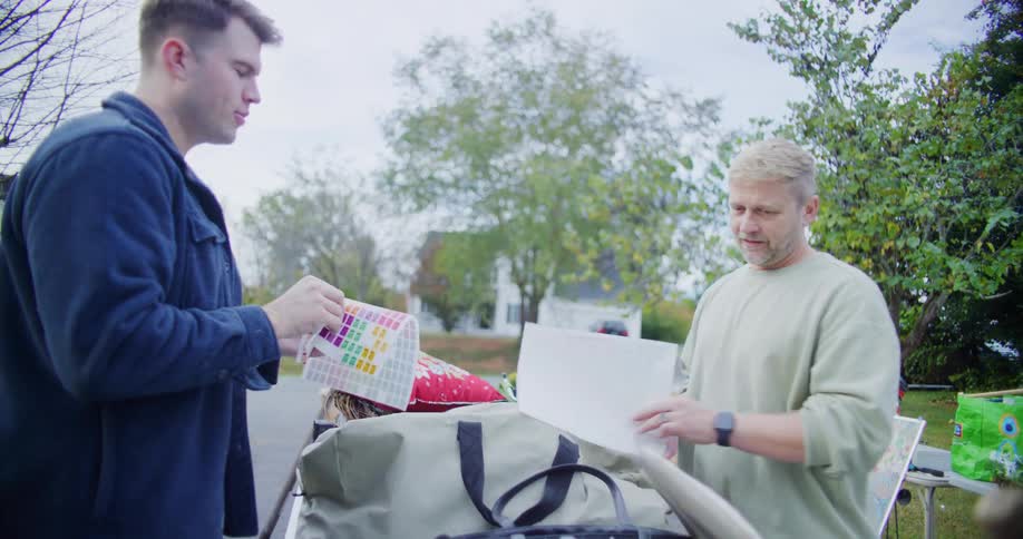 Men hold sticker sheets while organizing items on an outdoor table