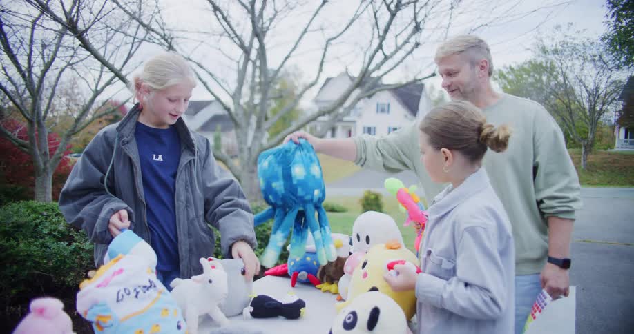A father smiles as he interacts with his two daughters who are looking at plush stuffed animals on a table at an outdoor yard sale in a suburban neighborhood