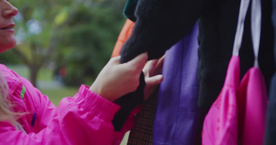 The video shows a closeup of a woman looking at and touching clothing that is hanging outdoors likely at a market or yard salea woman wearing a bright pink jacket with ruched cuffs is inspecting a rack of clothesher hand is shown touching and mov