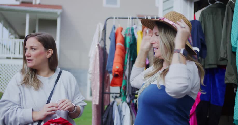 The video shows two women standing outdoors in front of a clothing rack likely at a yard sale or similar event the woman on the right tries on a straw hat while speaking animatedly to the woman on the left who is smiling and holding a small red garment