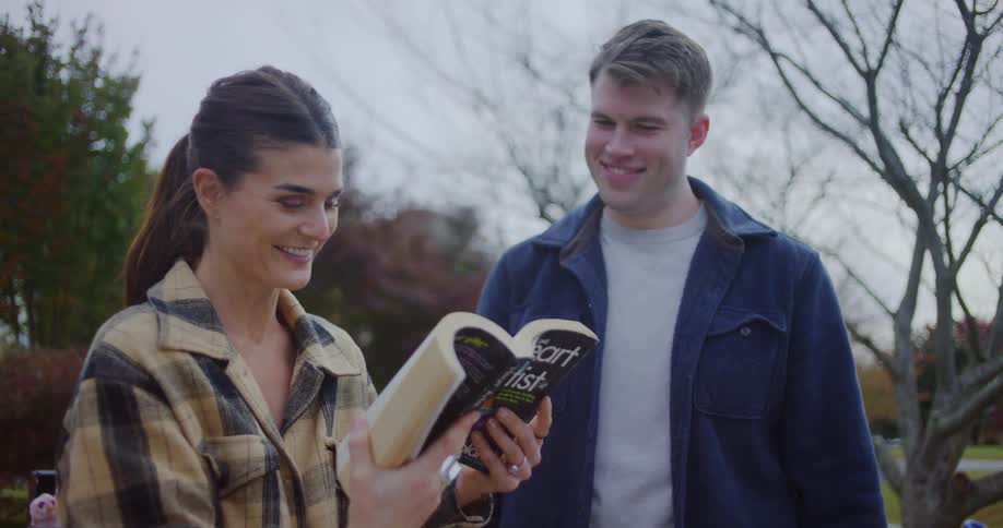 A woman wearing a plaid shirt holds up a book titled the heart of the artist and smiles while talking to a man in a blue jacket outdoors