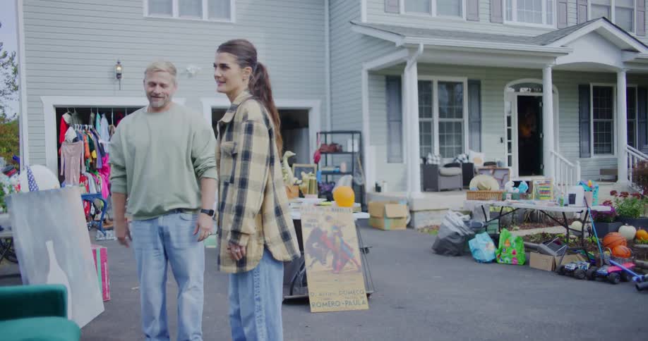 A smiling couple stands in the driveway of their suburban home shaking hands with a customer amidst a variety of furniture clothes and household items at a garage sale