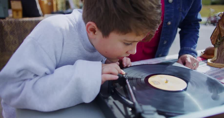 A child curiously watches a vinyl record spin while playing it fascinated by the motion beside him his mother stands outdoors during a midday garage sale sharing the moment together