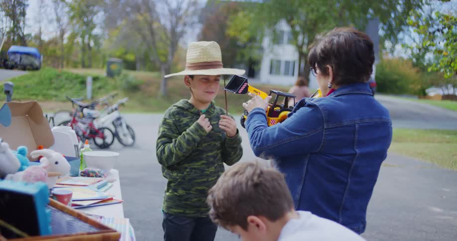 A mother and her two sons sort through items at an outdoor garage sale under the midday sun one boy playfully tries on a sun hat while the woman picks up a toy excavator