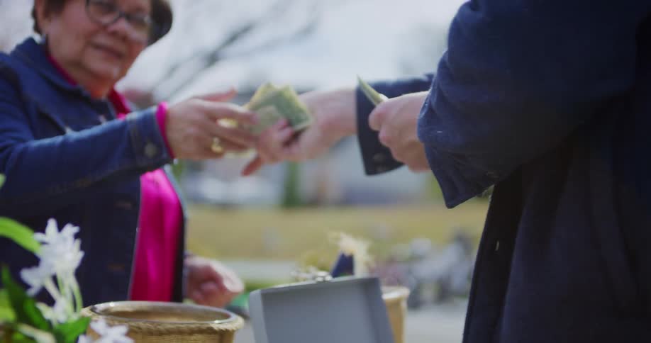 A man places money into an open cash box on a table next to a green thermos and straw basket then hands change to a smiling woman in a denim jacket and pink shirt outdoors