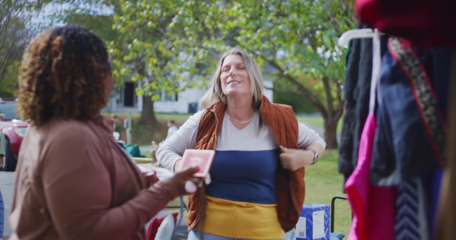 Woman tries on a coat at an outdoor thrift sale while another person watches