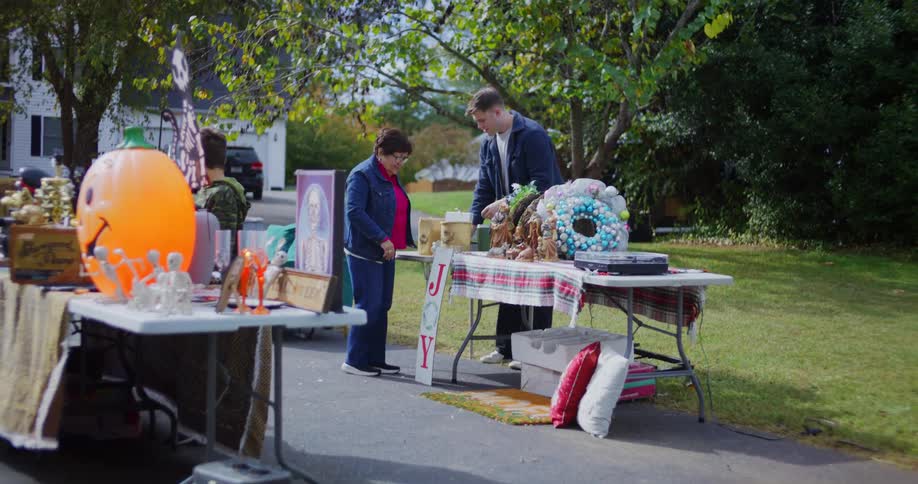 Two people browse holiday and decorative items on folding tables at a driveway yard sale
