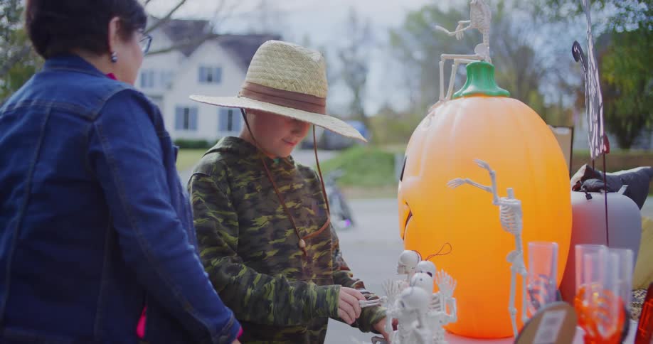 A woman and a boy examine halloween decorations beside a large orange pumpkin at a yard sale