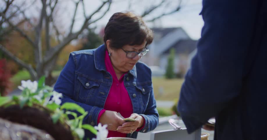 An older woman in a denim jacket and glasses smiles while paying at an outdoor yard sale in a suburban neighborhood