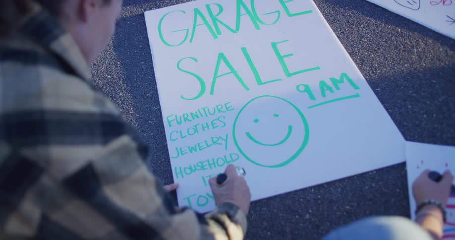 A young woman kneels outdoors at midday placing a sign on the ground with a marker in hand she carefully writes garage sale across it preparing for the event