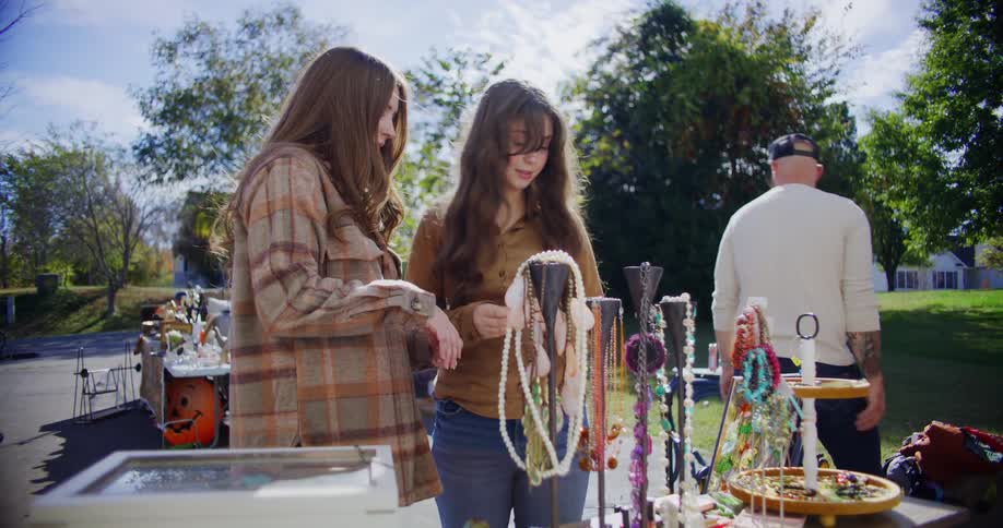 Two young women with long brown hair are looking at and touching jewelry displayed on stands at an outdoor yard sale on a bright sunny day