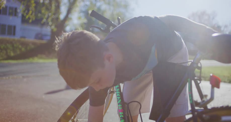 A young male in a dark shirt is focused on working on a bicycle outside on asphalt on a bright sunny day with a residential street visible