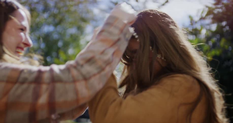 A young woman joyfully places a necklace around her teenage daughters neck both laughing with excitement under the bright midday sun outdoors