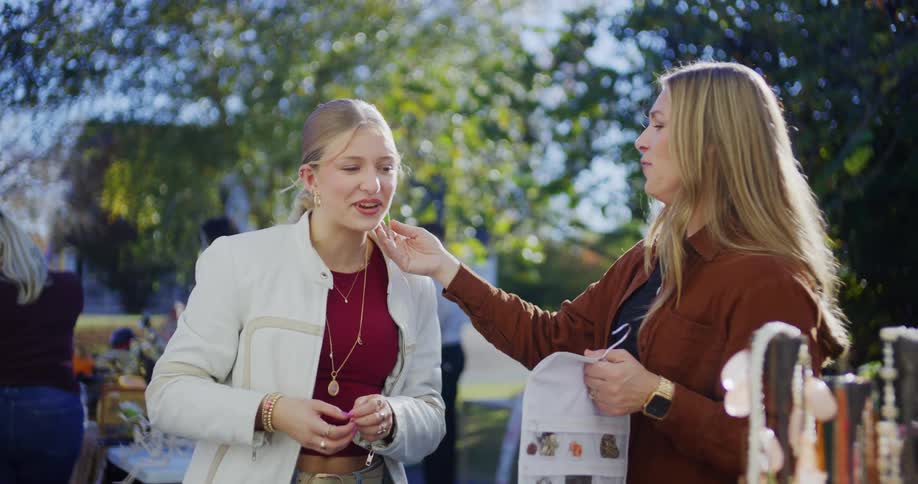 Two women chat at an outdoor market as one inspects the others earring