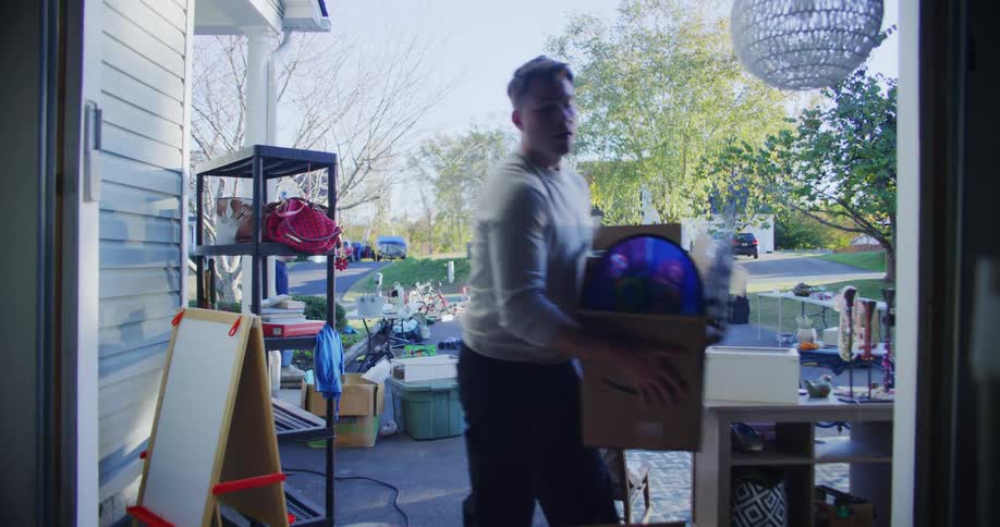 A young woman carries a box and hands it to a young man who takes it into the garage after setting it down he walks back to collect more items tidying up after a garage sale event