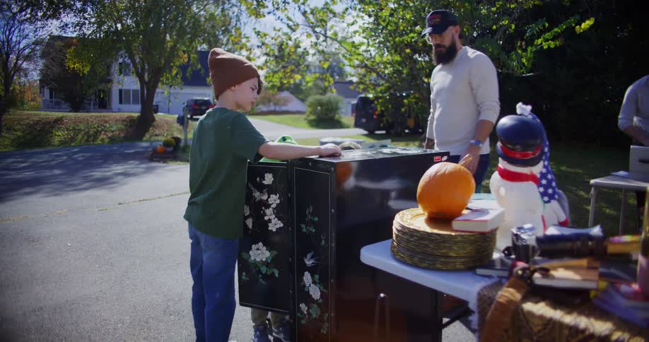 A family of two adults and two children stand around a black cabinet with floral decorations and other miscellaneous items at a yard sale in a suburban neighborhood on a sunny day