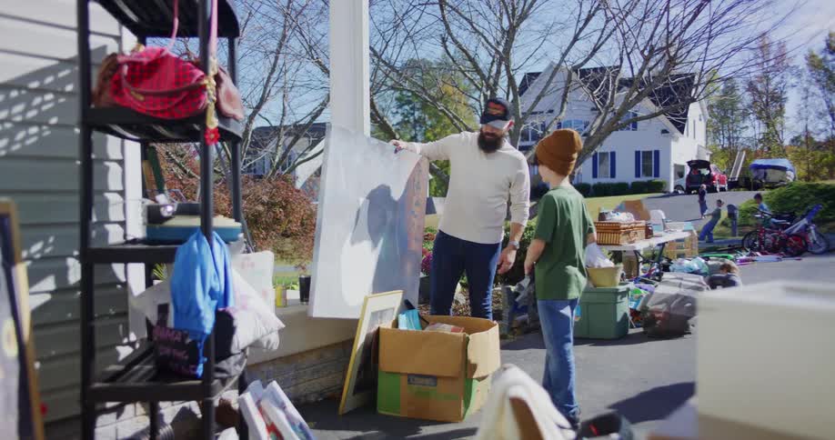 A bearded man in a white sweater and a baseball cap is standing on a porch during a yard sale talking to a young boy wearing a brown beanie the man picks up a large framed piece of artwork leaning against a pillar and shows it to the boy and a woman in