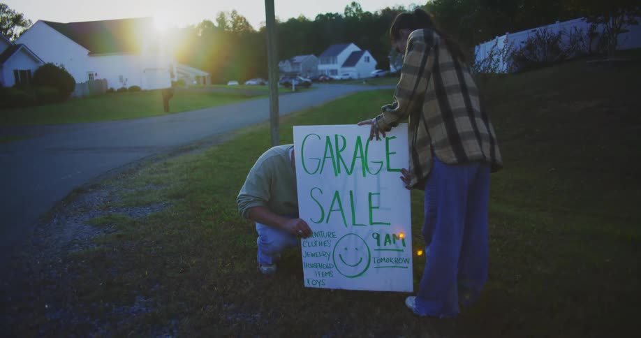 Young adult male and female putting a sign for a garagel sale in a lawn outdoors during sunrise