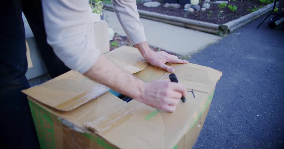 A person closes the flaps of a cardboard box filled with old items and carefully writes the word free across the top with a black marker