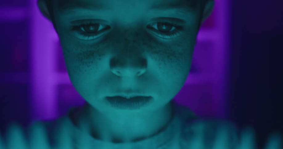 A young boy gazes down in wonder at a glowing blue book his face illuminated by the cool cyan light against a vibrant purple background as the camera slowly pushes in on his captivated expression