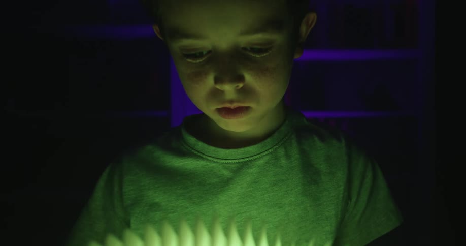A child stands in a dark room holding a light fixture the glow from it illuminates his face and clothes casting soft highlights against the surrounding shadows