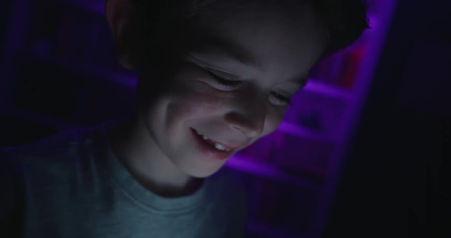A close view of a young boy smiling and laughing as his face is illuminated by a small screen or light source in a dark room with a purple illuminated bookshelf in the background