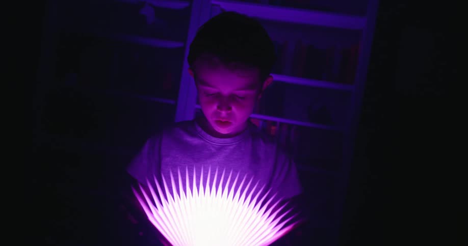 A close view of a young boy in a dark room illuminated by a bright magenta lightup paper book that is held open in his hands casting a glow on his surprised face