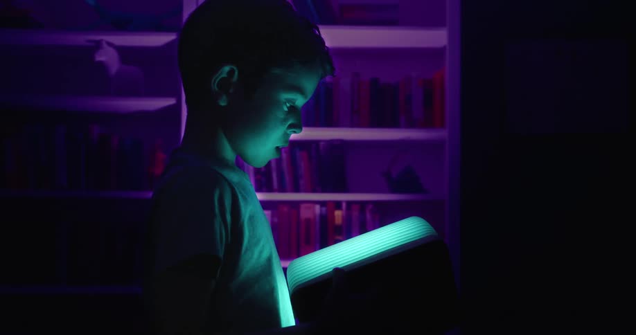 Boy standing in a dark room that contains bookcases is wowed by a glowing book