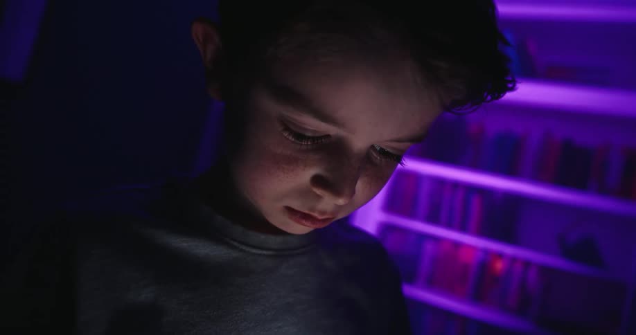 A close shot of a young boy looking down at a bright smartphone screen held in his hands which illuminates his face with cool light against a backdrop of a purple lit bookshelf in a dark room