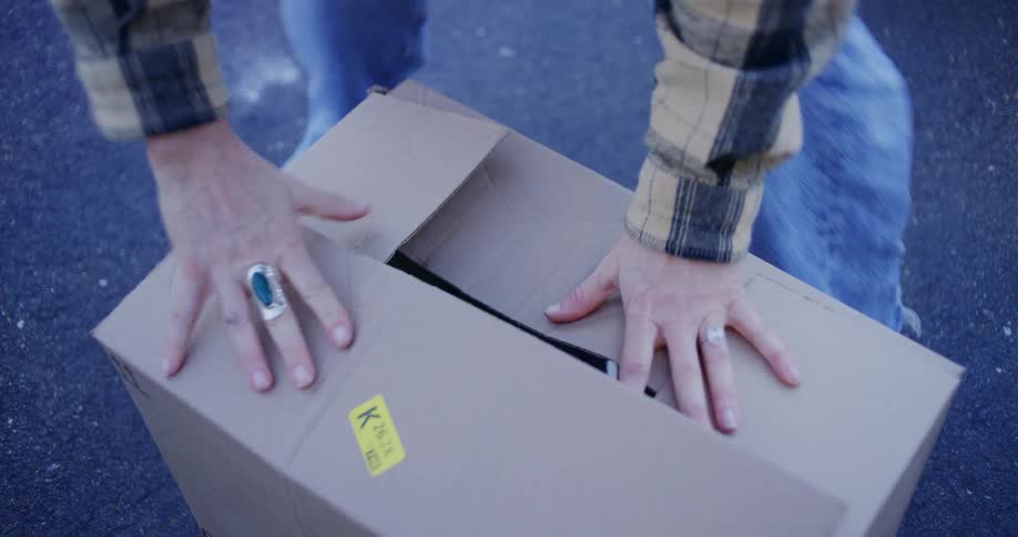 A person wearing a plaid shirt and jeans is kneeling down and carefully packing items into a cardboard box placing a patterned fabric storage bin inside first the box is then sealed with packing tape using a handheld dispenser indicating it is being pr
