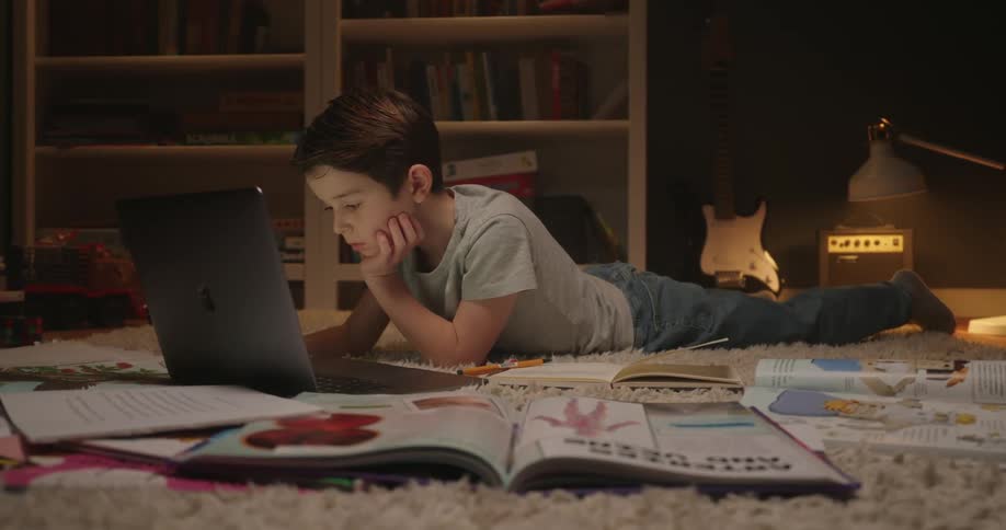 The boy is laying on the rug and typing on his laptop