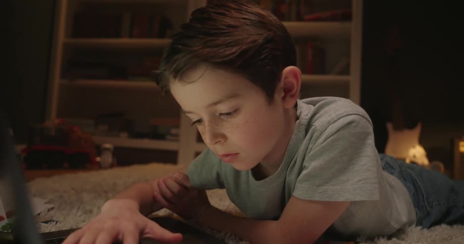 A young boy lies on a light carpet looking forward with concentration at a laptop in a dimly lit room with bookshelves in the background