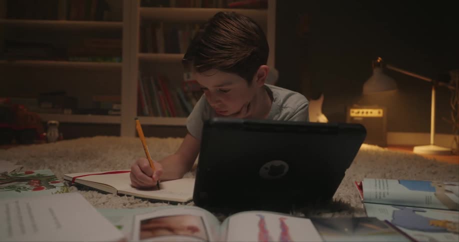 Child writes in a notebook while using a tablet resting on the rug
