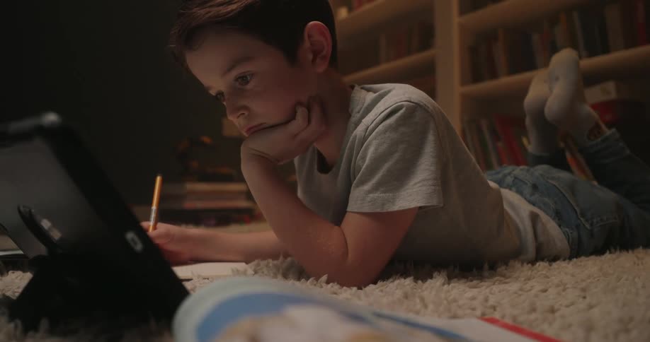 Child lies on the rug writing in a notebook while looking at a proppedup tablet