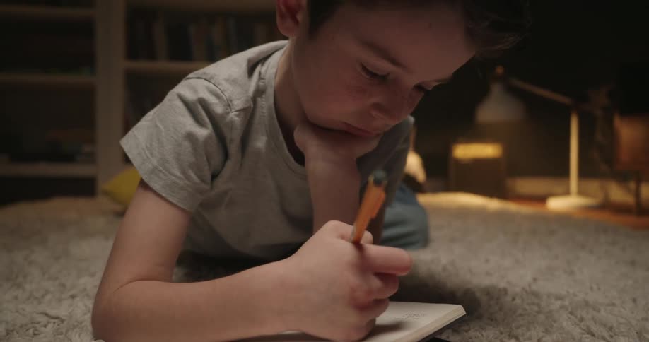 A young boy lies on a light carpet looking forward with concentration is writing in a notebook in a dimly lit room with bookshelves in the background