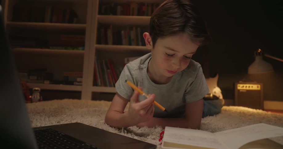 Child writes in a notebook while using a laptop resting on the rug