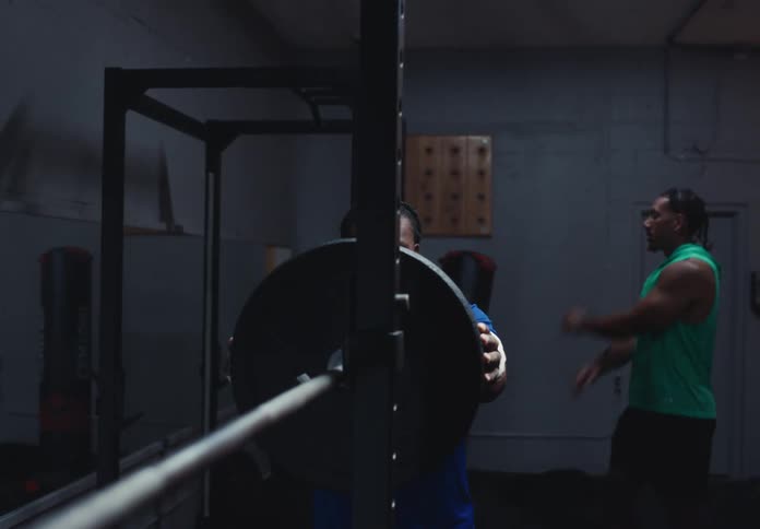 Person places a weight plate on a barbell while another prepares in the background
