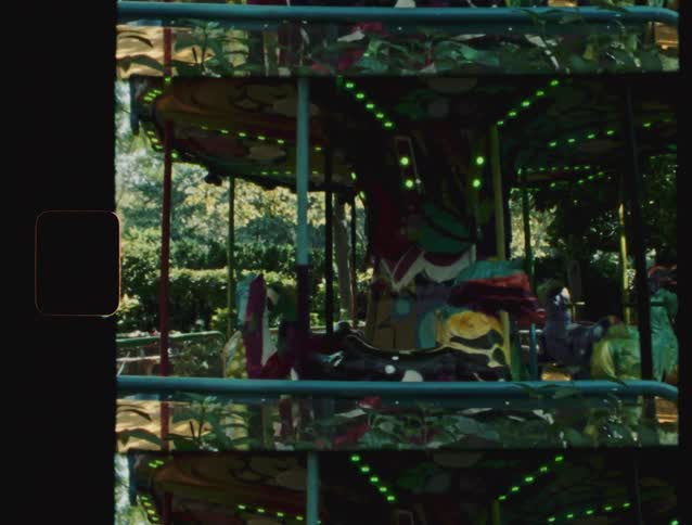 A colorful carousel in an amusement park slowly rotates in a lush green outdoor setting filmed on 16mm film stock with visible light leaks and film edges