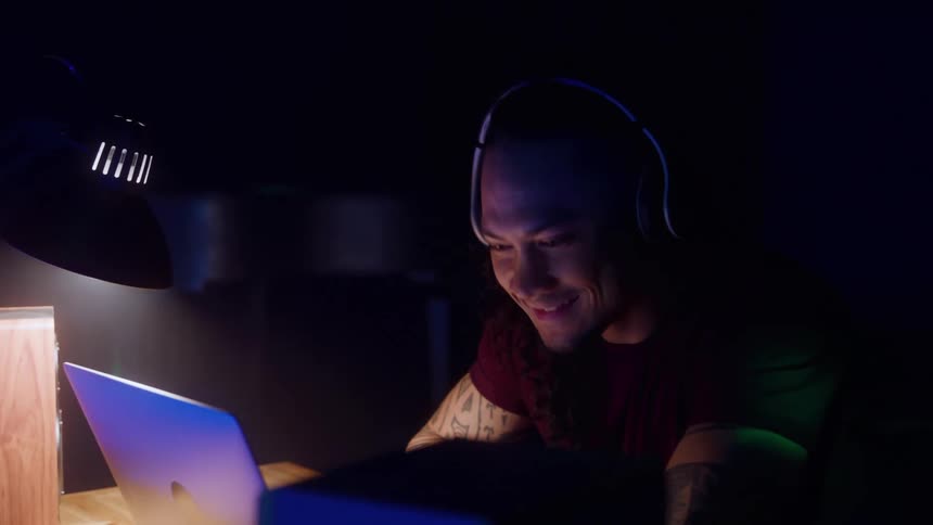 Man wearing headphones works on a laptop lit by a desk lamp